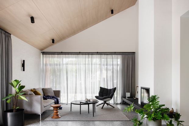 A refined contemporary living room with a light timber-lined sloping ceiling, white walls, full-height sheer curtains, and a large glazed opening. The room contains a pale upholstered sofa, dark lounge chair, round coffee tables, a textured grey rug, indoor plants, and a simple fireplace or framed wall feature. Black recessed or surface-mounted lighting and dark curtain hardware provide restrained contrast.