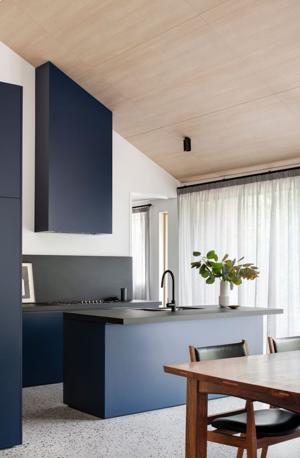 A refined open-plan kitchen and dining area with deep navy-blue cabinetry, a large island with an integrated black gooseneck tap and undermount sink, and a dark grey splashback. The kitchen opens to a dining zone with a warm timber table and dark leather chairs. Full-height sheer curtains provide abundant filtered daylight, while the pale timber-lined ceiling and terrazzo-style flooring add warmth and texture. A large leafy plant provides a natural accent.