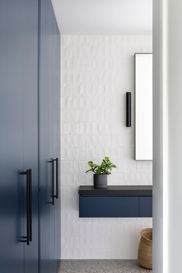 A compact, refined bathroom vanity area viewed through a doorway. Deep navy-blue built-in cabinetry with long matte-black pulls is set against a white textured rectangular-tile wall. A dark benchtop supports a small potted plant, with a white wall-mounted mirrored cabinet above. The floor is a speckled grey terrazzo-like finish, and a woven basket is partially visible at lower right. The palette is crisp white, navy, black, and grey with strong vertical lines and a clean contemporary feel.
