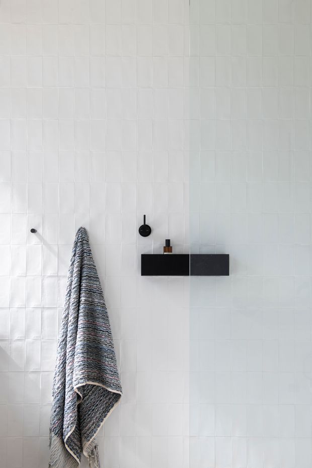 Minimalist bathroom wall clad in small white square tiles, featuring a black wall-mounted rectangular shelf or vanity element with a round black control, a small dark bottle, and a textured striped towel hanging from a simple hook. The composition is bright, restrained, and monochromatic with subtle tile variation and strong black accents.