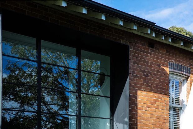 Close-up exterior view of a brick building facade with a large black-framed glazed opening and a traditional multi-pane window with white shutters. The dark reflective glazing mirrors surrounding trees and blue sky. A projecting roof edge with narrow clerestory-like openings is visible above, creating strong contrast between the warm brickwork, black framing, and bright reflections.