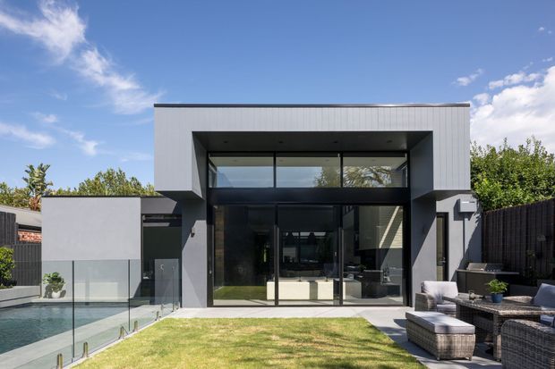 Modern two-storey residential facade viewed from the backyard, with a crisp rectilinear form, dark grey and light grey exterior finishes, expansive black-framed glazing, and a recessed upper-level opening. A glass-edged swimming pool, lawn, and outdoor lounge area occupy the foreground.