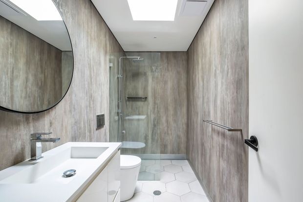A bright, contemporary bathroom featuring full-height wood-look wall panels, a white floating vanity with integrated basin, a large rounded mirror, wall-hung toilet, frameless glass shower enclosure, and white hexagonal floor tiles. Skylights or recessed ceiling lights provide strong natural-looking illumination.