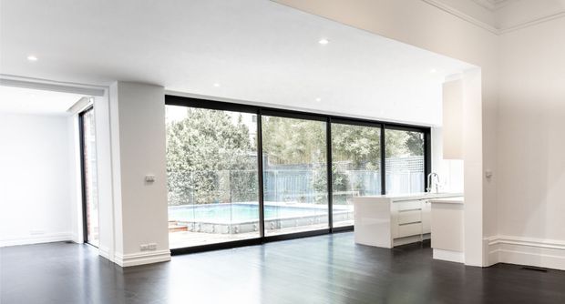 Bright open-plan interior with a contemporary white kitchen at the right, dark timber-look flooring, recessed ceiling lights, decorative cornices and skirting, and expansive black-framed floor-to-ceiling glazing overlooking a swimming pool and garden. An adjoining open room is visible to the left.