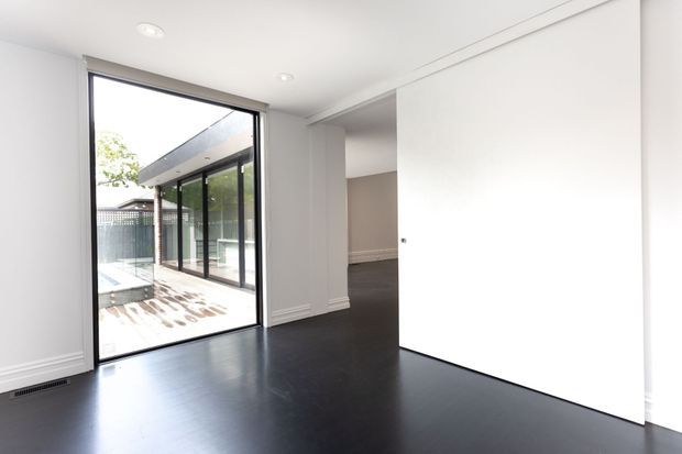 Bright, minimal contemporary interior with dark timber flooring, white walls and recessed ceiling lights. Full-height glazing and a sliding glass door open onto a paved outdoor area, while a large white sliding panel conceals an adjoining space.