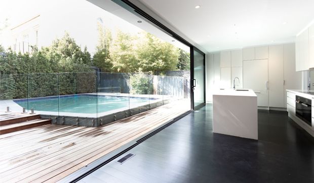 A bright contemporary kitchen opening directly onto a timber deck and swimming pool through expansive black-framed sliding glass doors. The kitchen features a white island with integrated sink, streamlined white floor-to-ceiling cabinetry, dark timber flooring, and a clean minimalist fit-out. The outdoor pool and deck are clearly visible beyond the glazing.
