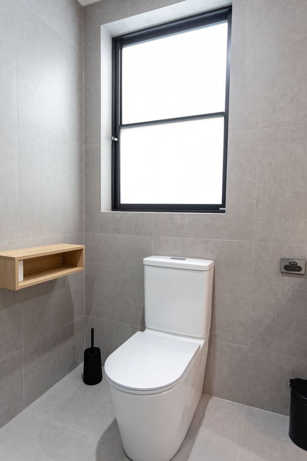 A compact contemporary bathroom featuring a white toilet, large-format grey wall and floor tiles, a black-framed frosted or overexposed window, and a simple light timber wall shelf. Black accessories and hardware provide contrast against the neutral finishes.
