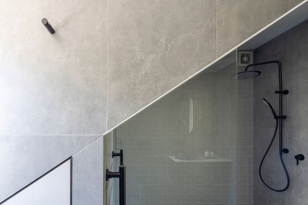 Close-up of a contemporary bathroom shower area with large grey concrete-look wall tiles, a frameless glass shower screen, and matte black shower hardware including an overhead rain shower and handheld shower. A diagonal ceiling or wall line creates a distinctive geometric composition, with a recessed shelf visible through the glass.