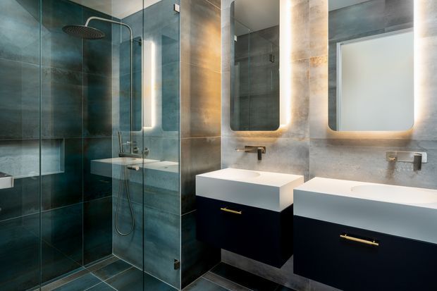 Contemporary bathroom featuring a glass-enclosed walk-in shower, dual wall-mounted vanity units, illuminated rounded-rectangle mirrors, dark teal-grey wall and floor tiles, pale stone-look surfaces, and warm brass tapware and handles. The space has a refined, high-contrast, softly lit appearance.