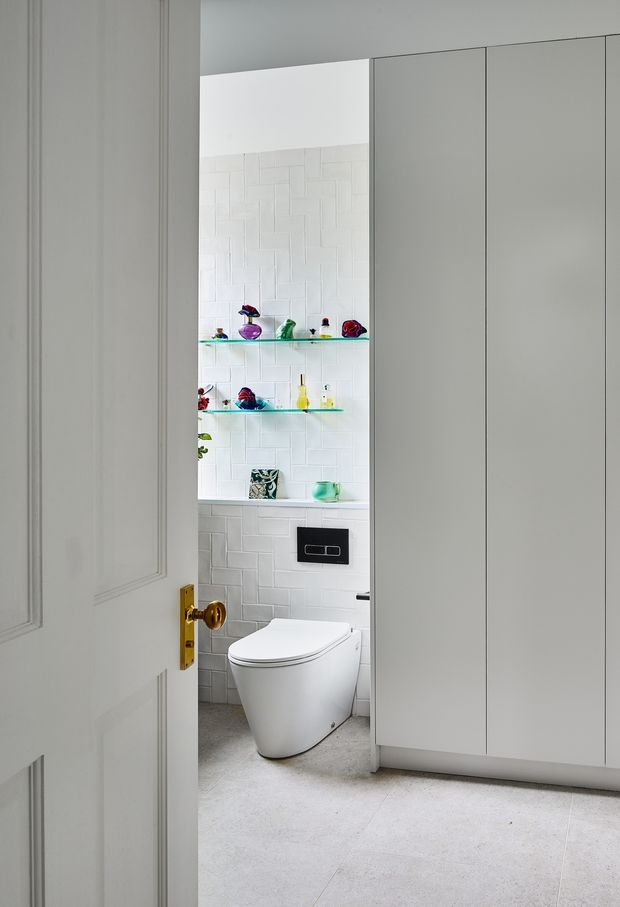 A bright, compact contemporary bathroom viewed through a partially open paneled door. The room features a wall-hung toilet with a black flush plate, white textured rectangular wall tiles, pale tiled flooring, open glass display shelves with colourful decorative objects, and tall minimalist white built-in cabinetry.