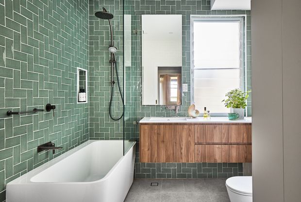 Contemporary bathroom featuring a white freestanding bathtub with wall-mounted black fixtures and a glass shower screen. The walls are clad in dark sage-green rectangular tiles arranged in a herringbone pattern, complemented by a timber vanity with integrated drawers, a white basin, mirrored cabinet, and a bright window. Grey floor tiles and restrained accessories create a refined, natural-toned scheme.