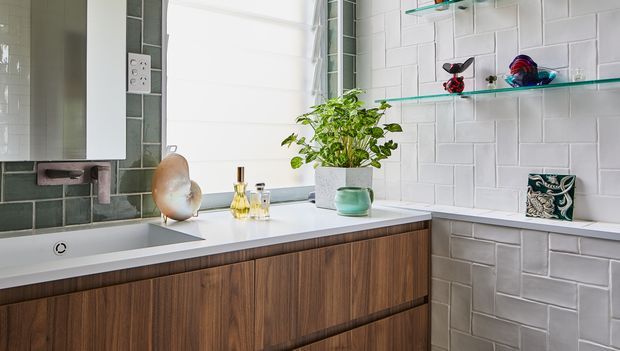 Bright contemporary bathroom vanity with a long white benchtop, warm timber-grain floating cabinetry, and a freestanding white basin at left. The walls are finished in contrasting dark grey and white glazed rectangular tiles, with open glass shelves displaying small decorative objects and a potted plant. A wall-mounted mirrored cabinet and electrical outlet are visible, creating a clean, refined fit-out with natural warmth.