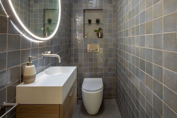 Compact contemporary bathroom with floor-to-ceiling blue-grey square wall tiles, matching grey floor tiles, a white integrated basin on a floating wood-grain vanity, wall-mounted brushed metal tapware, and a compact toilet. A large rounded backlit mirror and recessed display niche with decorative plants add soft illumination and detail.