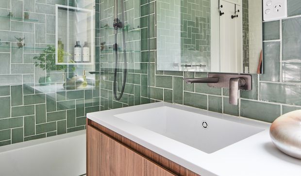 Close-up view of a contemporary bathroom featuring a white integrated bathtub with timber-look vanity cabinetry, a wall-mounted brushed metal tap, and a large mirrored cabinet. The walls are clad in glossy green rectangular tiles arranged in a vertical-and-horizontal stacked pattern, with a glazed shower enclosure and recessed shelving visible to the left. The palette is fresh and refined, combining green tile, white sanitaryware and warm wood tones.