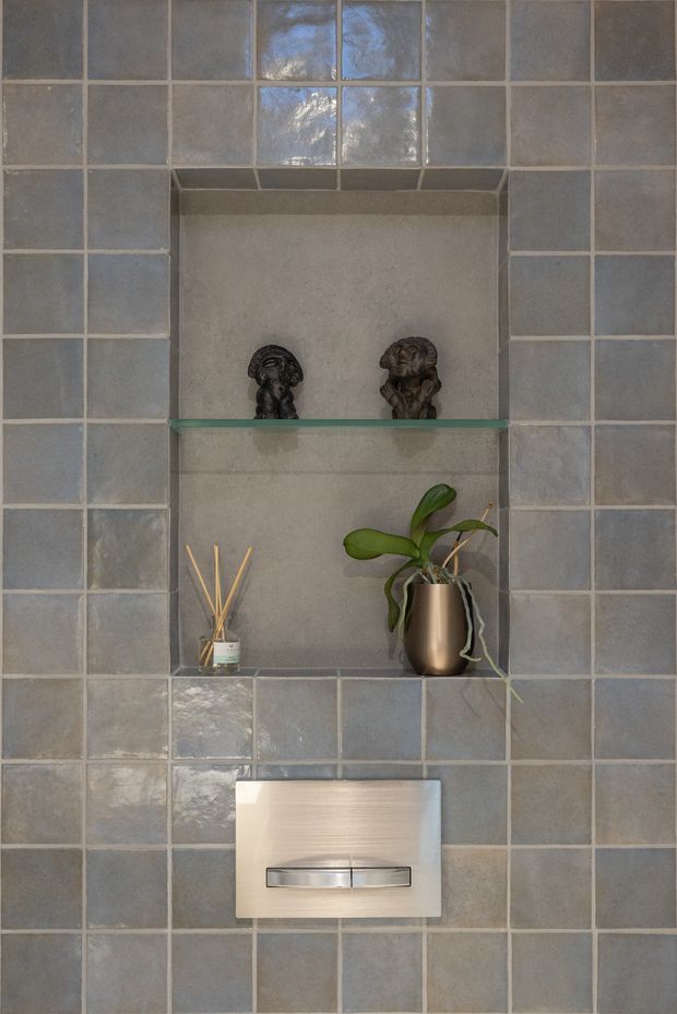 Close-up view of a tiled bathroom wall featuring a recessed display niche with a glass shelf, small decorative sculptures, a reed diffuser, and a potted plant. Below the niche is a flush-mounted wall control or fixture. The wall is finished in square, blue-grey glazed tiles with light grout, creating a muted and reflective surface.