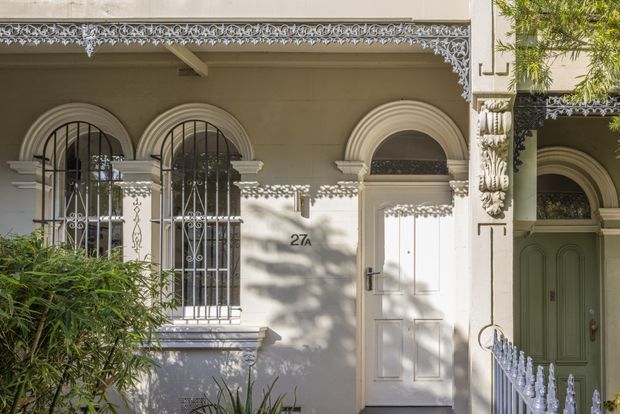 Street-facing facade of a historic terrace or townhouse, featuring cream rendered walls, arched windows and door surrounds, decorative wrought-iron window grilles, ornate lacework frieze, sculptural column detailing, and contrasting white and pale green entry doors. Mature planting partially frames the frontage, with strong dappled sunlight creating patterned shadows.