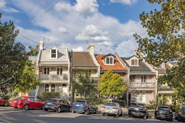 Street-level exterior view of a row of attached and closely spaced heritage-style urban houses. The façades feature pitched gabled roofs, dormer windows, rendered walls, decorative verandas and balconies with ornate balustrades, chimneys, and varied roof materials. Mature street trees and parked cars line the foreground.