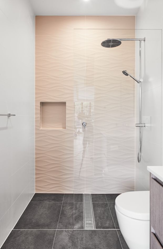A bright, contemporary walk-in shower within a bathroom. The shower features a frameless glass screen, dual chrome showerheads with handheld wand, a recessed wall niche, pale pink-beige three-dimensional wave-textured wall tiles, white side wall tiles, and dark grey floor tiles with a linear stainless-steel drain. Part of a white toilet and timber-toned vanity are visible at the right edge.