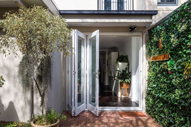 Exterior view of a contemporary residential entry courtyard, with a glazed bi-fold doorway opening into a bright hallway. The entrance is framed by pale rendered walls, dark timber flooring visible inside, a planted green wall with a small address plaque, potted greenery, and a tiled or paved threshold area.