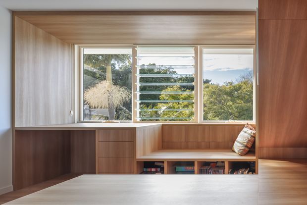 A compact, highly integrated timber-lined interior with built-in cabinetry, shelving, and a broad window seat beneath three louvred windows. Warm wood joinery frames views of greenery outside, while a partial upholstered cushion and books add residential character. The space appears to function as a study or living-room reading nook, with clean contemporary detailing and abundant natural light.