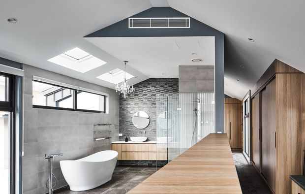 A spacious contemporary bathroom with a freestanding white bathtub, walk-in glass shower, double vanity, wall-mounted fixtures, and extensive built-in timber cabinetry. Large-format grey wall and floor tiles contrast with a dark patterned feature wall behind the vanity. The sloped ceiling includes skylights and a high-level ventilation grille, while a glazed opening and doorway provide abundant natural light.