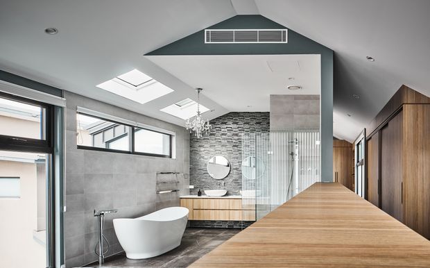 Contemporary bathroom with a freestanding white bathtub, wall-mounted vanity, large timber island-style counter, textured grey stone wall, frameless glass shower enclosure, skylights, and dark timber built-in cabinetry. The room has a high vaulted ceiling with an integrated ventilation grille and a refined neutral palette.