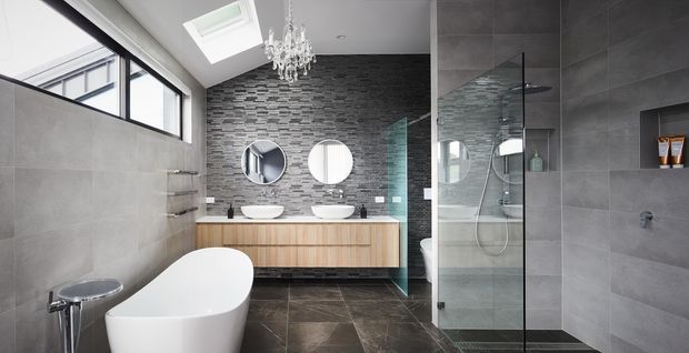 Contemporary, spacious bathroom with a freestanding white bathtub, double vessel basins on a timber vanity, large-format grey wall tiles, dark tiled flooring, a textured dark feature wall, and a frameless glass shower enclosure. High windows, a skylight, and a crystal chandelier provide natural and decorative lighting.