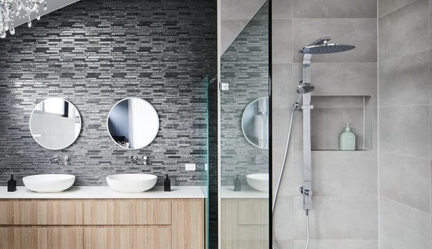 Contemporary bathroom with a double vanity featuring white vessel basins, round mirrors, light timber cabinetry, and dark textured stone mosaic wall cladding. A frameless glass shower enclosure with a chrome rain shower and recessed wall niche occupies the right side. The space has a refined monochromatic palette with warm timber accents and a decorative chandelier partially visible at the top left.