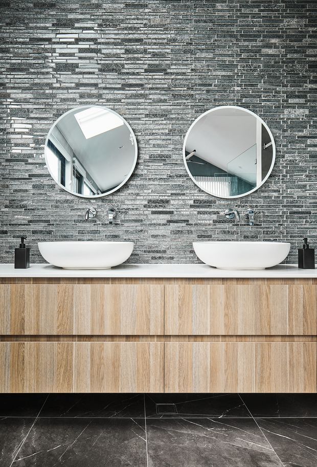 A contemporary double-vanity bathroom featuring two white vessel basins on a long light-wood floating vanity. The wall is clad in narrow stacked grey stone-look tiles, with round frameless mirrors, wall-mounted chrome taps, black dispensers, and large dark marble-look floor tiles. Mirror reflections show a bright skylight and part of the surrounding interior.