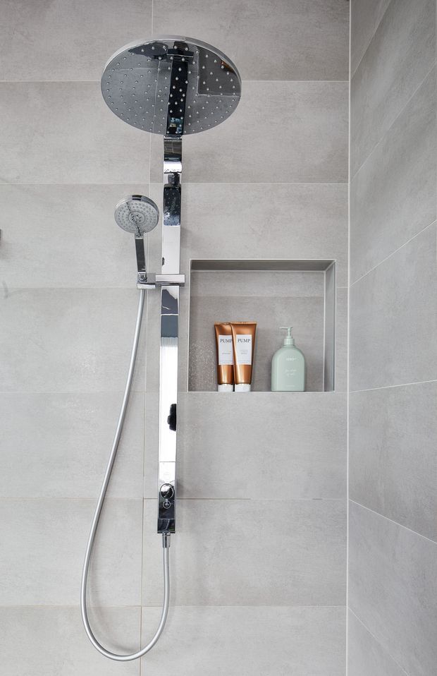 Close-up of a contemporary shower enclosure with large-format light grey wall tiles, a chrome thermostatic shower column, overhead rain shower, handheld shower, and a recessed wall niche containing toiletries.