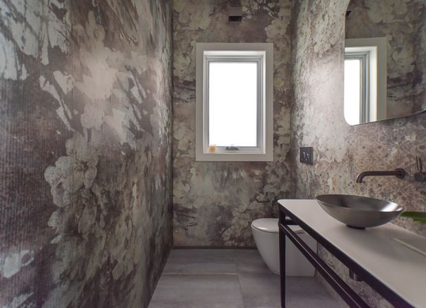 A compact contemporary bathroom with a wall-mounted basin on a dark metal-framed vanity, a vessel sink, toilet, wall-mounted tapware, mirror, and small window. The walls feature a dramatic distressed, mottled botanical-style finish in grey, taupe, brown, and muted green tones, contrasted with large-format grey floor tiles and white sanitaryware.