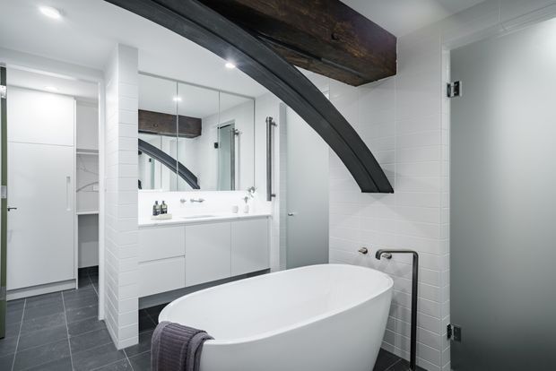 Bright contemporary bathroom with a freestanding white bathtub, wall-mounted white vanity, large mirror, glass shower enclosure, white tiled walls, dark tiled flooring, and a dramatic exposed dark timber beam intersecting a curved dark structural element. A wardrobe or storage area is visible through the opening at left.