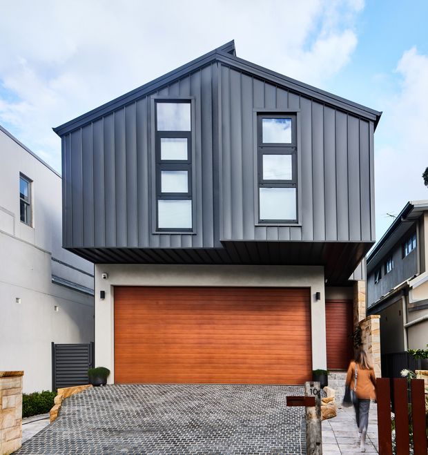 Front exterior view of a narrow multi-storey contemporary home in an urban setting. The upper level is clad in dark grey vertical standing-seam metal with a steep gabled roof and tall stacked windows. A projecting upper storey shelters a ground-level garage with a warm timber sectional door. The entry driveway is paved in a dark patterned finish, with neighbouring attached or closely spaced homes visible on both sides.