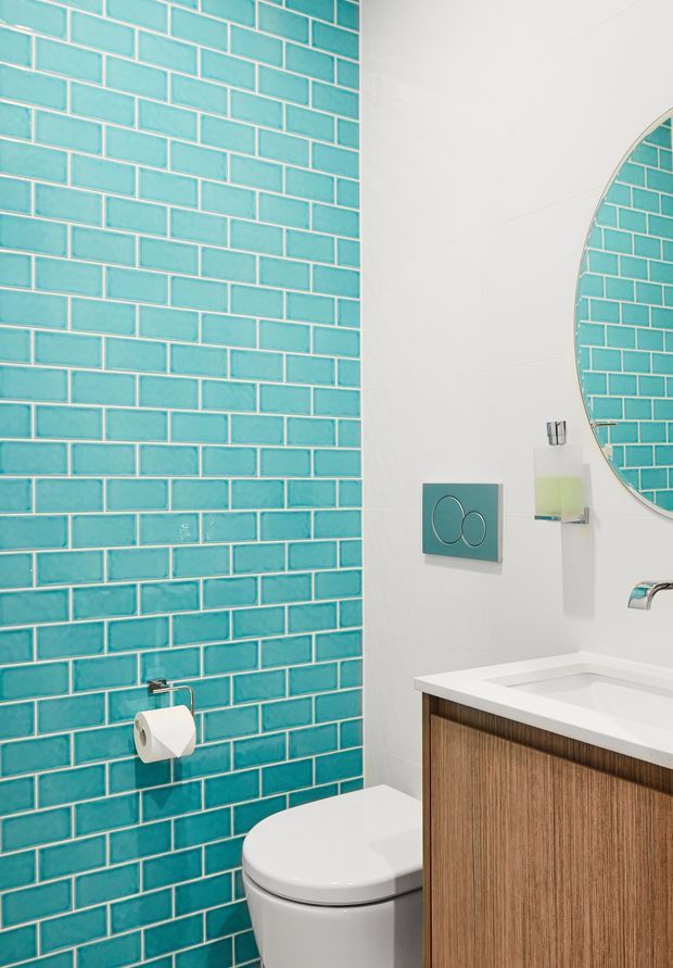 A compact, contemporary bathroom featuring a vivid turquoise subway-tile feature wall, white painted walls, a wall-mounted dual-flush plate, round mirror, white integrated basin, timber-toned vanity cabinetry, and a white toilet. Chrome fixtures and a pale soap dispenser add functional details.