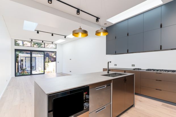 Bright contemporary kitchen with a large central island, integrated appliances, extensive blue-grey and bronze-toned cabinetry, pale timber flooring, pendant lights, track lighting, skylights, and glazed doors opening to an outdoor area.