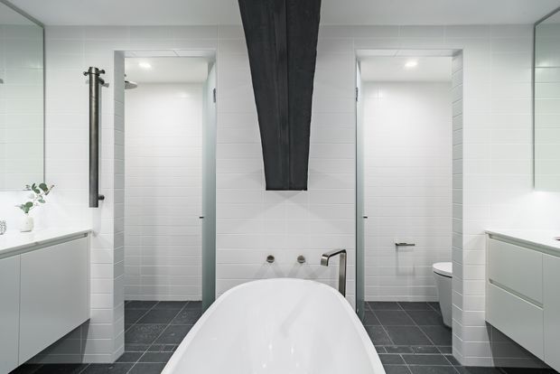 Bright contemporary bathroom with a freestanding white bathtub in the foreground, positioned between two tiled shower and toilet areas. White horizontal wall tiles, dark grey floor tiles, pale grey floating vanities, mirrored cabinetry, and a dramatic dark vertical feature above the bath create a clean, high-contrast scheme.