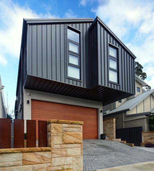 Street-facing view of a contemporary multi-storey residence with a strongly projecting upper volume clad in dark vertical metal panels. The facade features narrow stacked windows, a warm timber garage door, sandstone masonry walls, and a broad paved driveway. The composition emphasizes the modern angular building form against a partly cloudy sky, with a neighboring pitched-roof home visible to the side.