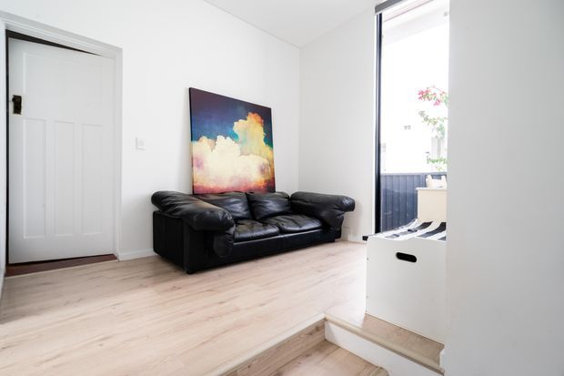 Bright, compact living room with white walls, pale timber-look flooring, a black leather sofa, and a colourful cloud artwork. A short step-down leads toward a bright glazed opening or balcony area, while a white paneled door sits to the left.