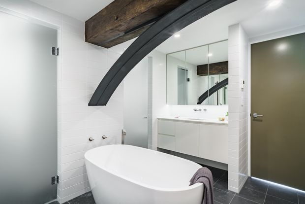 Contemporary bathroom with a freestanding white bathtub, double vanity, mirrored wall, white tiled walls, dark tiled floor, and a distinctive exposed timber beam paired with a curved dark structural element. A frosted glass door and recessed ceiling lighting complete the clean, high-contrast space.