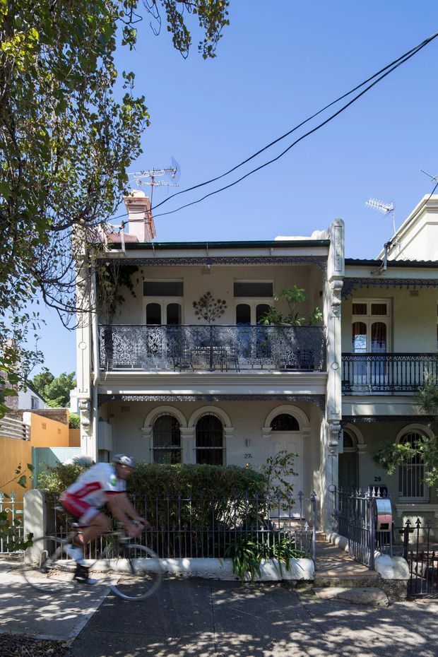 Street-facing view of a narrow, attached two-storey terrace house with an ornate historic facade. The building features arched ground-floor openings, decorative moulded trim, lace-like metal balcony balustrades, slender columns, a parapet roofline, and a small planted front garden behind a metal fence. A neighbouring terrace is visible to the right, with overhead utility wires and a cyclist in the foreground.