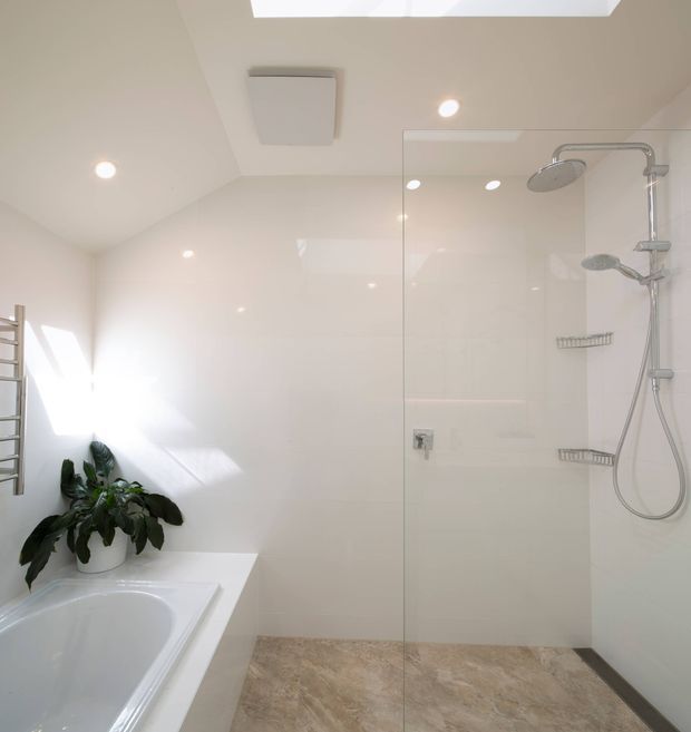Bright contemporary bathroom with a large frameless glass walk-in shower, dual showerheads and chrome fixtures, a white built-in bathtub, pale glossy wall surfaces, beige stone-look floor tiles, recessed lighting, a skylight, and a potted plant. A heated towel rail is visible at the left.