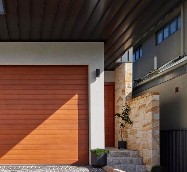 A close architectural view of a covered residential entry and garage facade. The scene features a large horizontal timber-toned sectional garage door, a matching pedestrian door, white rendered walls, dark metal ceiling panels and window frames, pale stacked-stone cladding, and stone steps leading toward the entry. Strong sunlight creates a diagonal shadow across the garage door, highlighting the warm contemporary material palette.