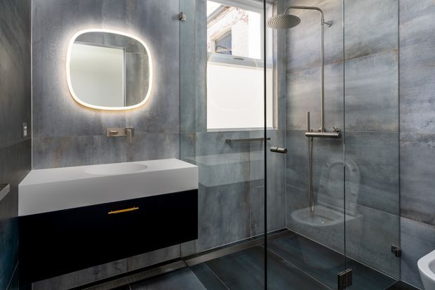 Contemporary bathroom with large grey concrete-look wall tiles, dark floor tiles, a floating black vanity with white integrated basin, and a frameless glass walk-in shower. A rounded illuminated mirror and exposed brushed metal shower fittings add modern functional detailing. A partial freestanding bath is visible at the right edge.