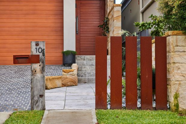 Residential front entry viewed from the street, featuring a warm timber-clad garage or entry door, a paved driveway with small dark interlocking pavers, pale concrete or stone stepping slabs, sandstone retaining and boundary walls, and a contemporary rust-coloured vertical metal fence. A timber-and-metal letterbox marked 10 stands beside the path, with planted greenery around the entry.
