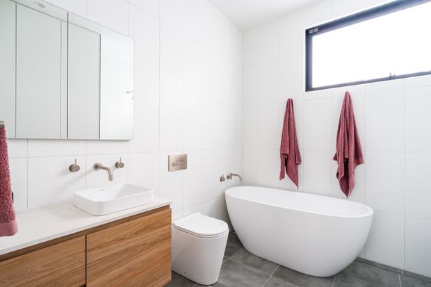 Bright contemporary bathroom with large white wall tiles, grey tiled flooring, a wall-mounted timber vanity with white vessel basin, mirrored storage cabinetry, wall-mounted chrome tapware, a freestanding white oval bathtub, and a white toilet. A high horizontal window provides natural light, while burgundy towels add colour contrast.