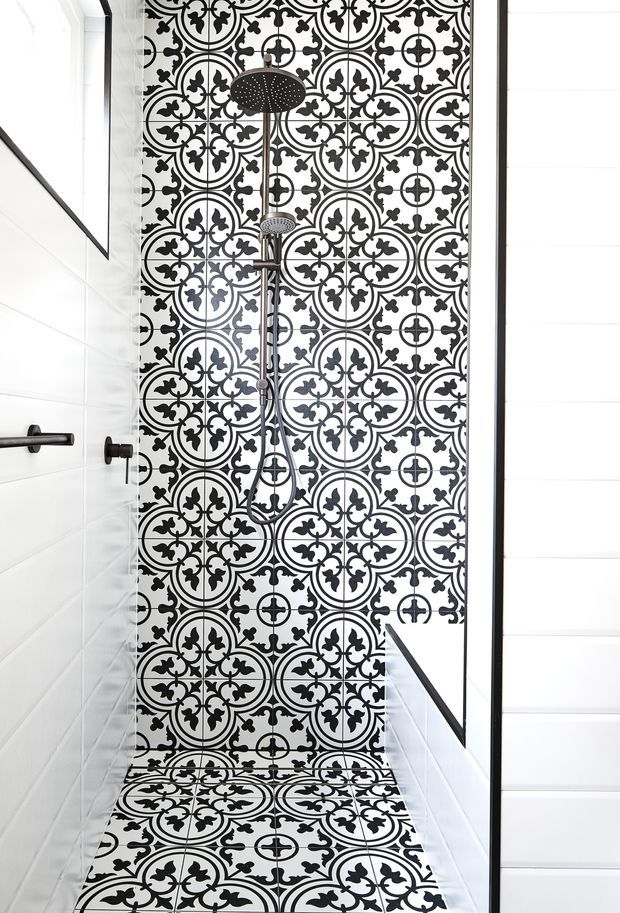 Bright, narrow bathroom shower enclosure featuring a bold black-and-white patterned tile feature wall that continues onto the shower floor. A black rain shower and handheld shower set are mounted centrally, with white wall tiles, dark fixtures, and a black-framed glass shower screen.