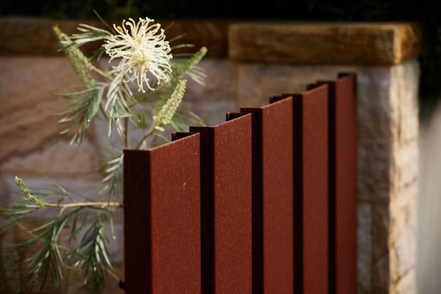 Close-up exterior detail of a dark reddish-brown vertical slatted panel or screen set against a rustic stone and timber backdrop, with native foliage and a pale flower beside it. The image focuses on the panel's textured finish and repeating geometric profile.