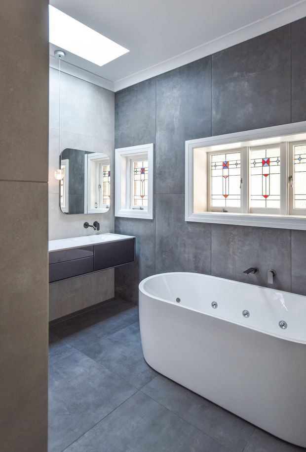 A contemporary bathroom with a freestanding white spa bath, floating dark vanity, wall-mounted basin tapware, large-format grey tiled walls and floor, and decorative stained-glass windows. A skylight provides natural light, while a mirrored cabinet is mounted above the vanity.