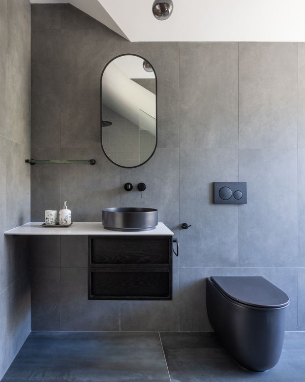 Contemporary compact bathroom featuring large-format grey wall and floor tiles, a floating dark timber vanity with a white benchtop, round dark metal vessel basin, wall-mounted black toilet, frameless arched mirror, and minimalist black fixtures. The room has a restrained monochromatic palette and clean-lined built-in detailing.
