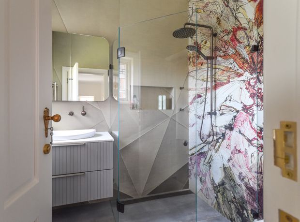 A contemporary bathroom viewed through an open door, featuring a floating grey vanity with a white basin, rounded wall mirror, wall-mounted taps, and a frameless glass walk-in shower. The shower has dark fittings and geometric grey wall tiles, while a bold abstract red, black, and white patterned feature wall adds visual contrast. The room has a refined, artistic and modern character.